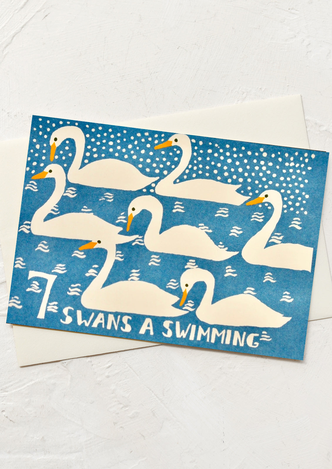 A square greeting card with a blue background and white illustrated swans, with the text '7 SWANS A SWIMMING' at the bottom, shown on a white textured surface with a white envelope behind it.