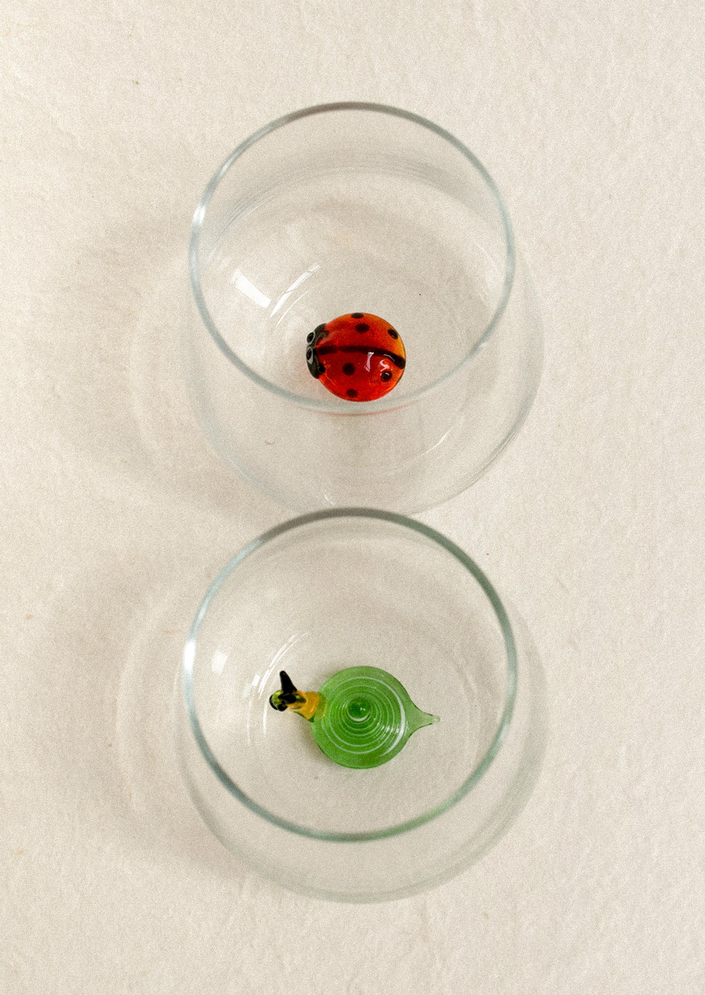 Two clear glasses, each containing a small decorative glass figure: a red ladybug in the top glass and a green snail form in the bottom style.