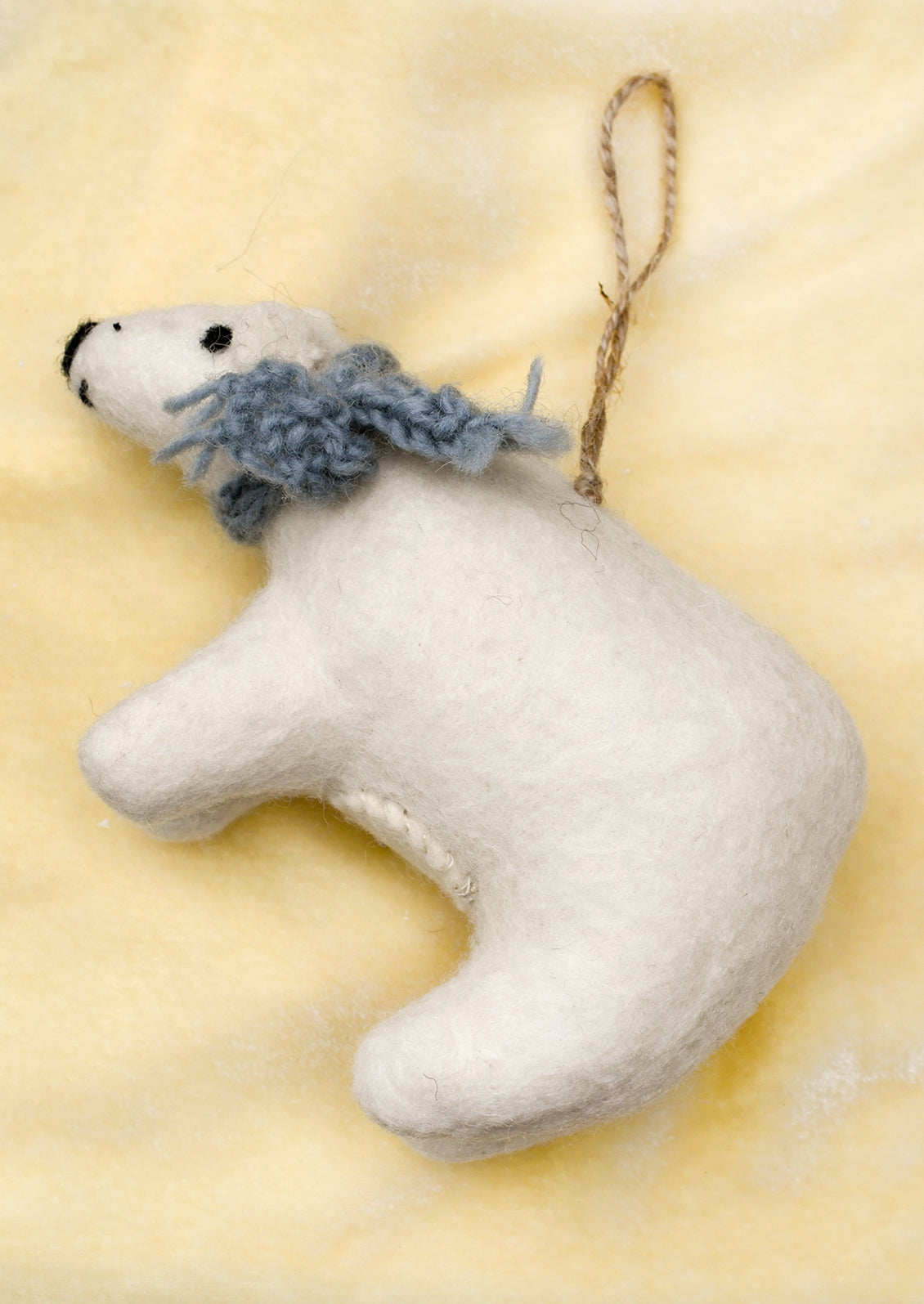 A white felt polar bear Christmas ornament with a knitted blue-gray scarf and a brown jute hanging loop.