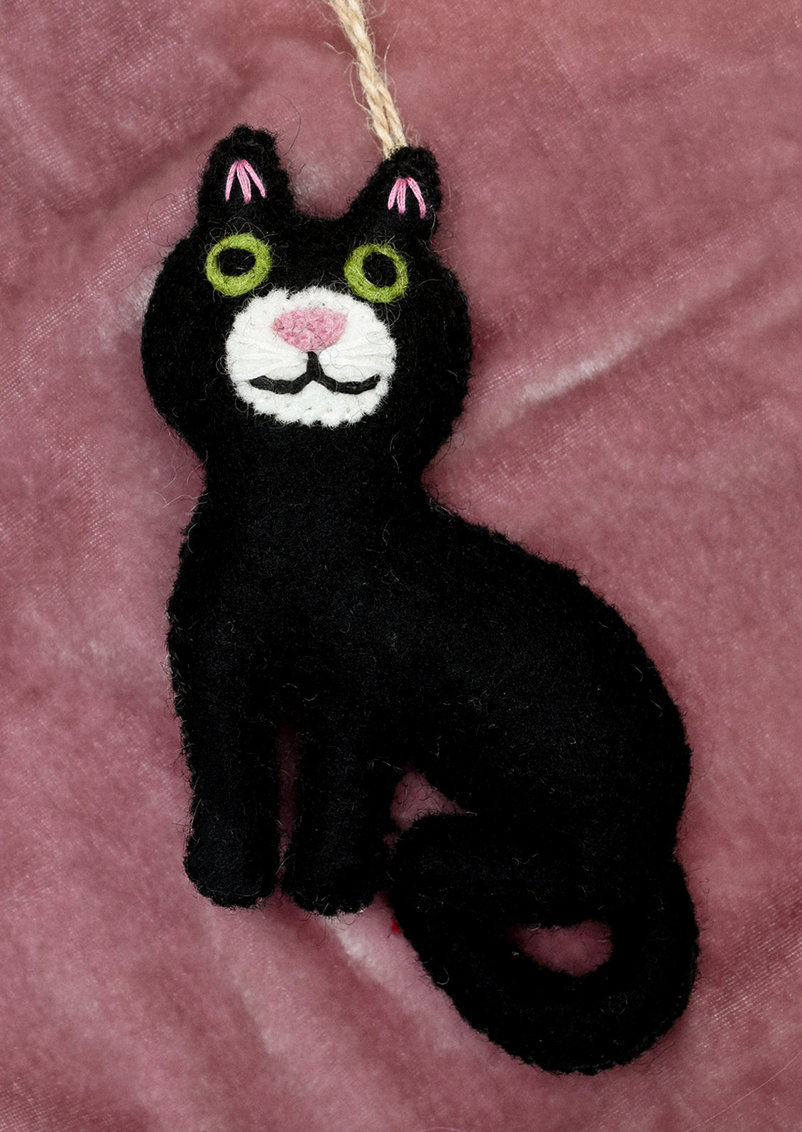 A black felt cat ornament with embroidered green eyes, pink nose and ear accents, and a white chin, hanging from a tan string against a white fabric background.
