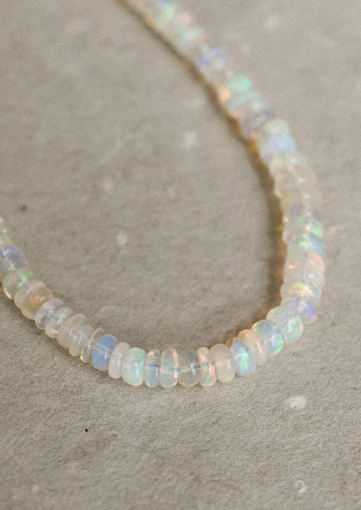 A beaded necklace made of small, irregular opal-like beads that shimmer with iridescent colors of white, pale blue, and soft yellow, laid out on a textured beige surface.