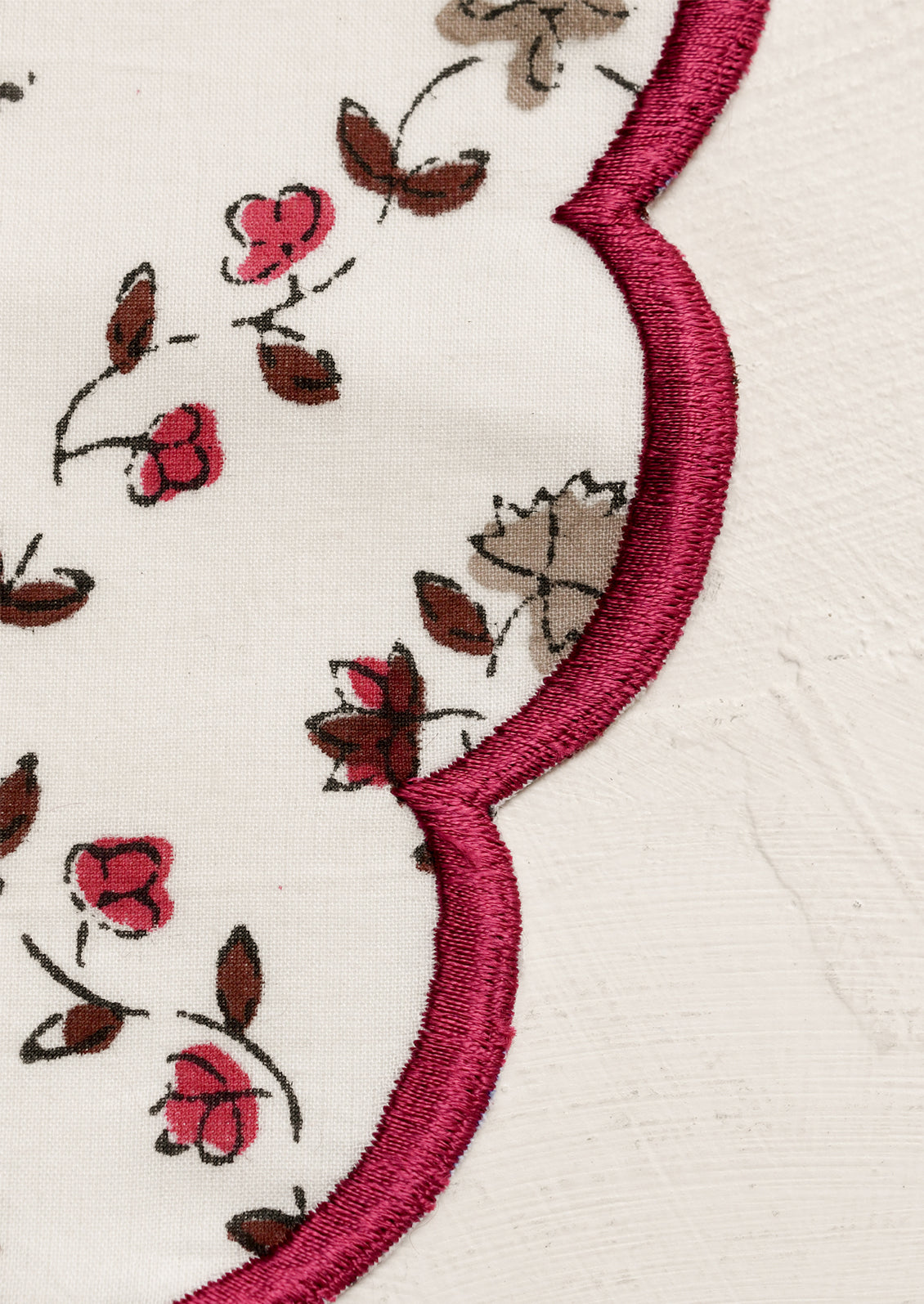 A round scalloped placemat in white with tan and burgundy floral print and embroidered burgundy trim.