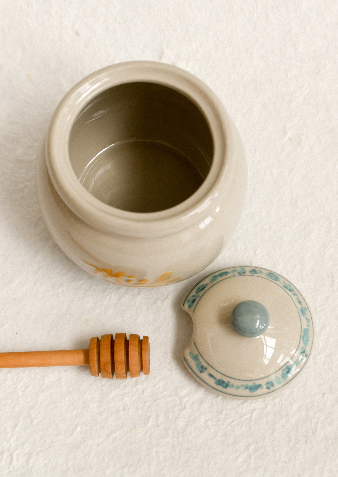 A stoneware honey jar with "Miel" written in yellow script, with blue floral stripe trim.