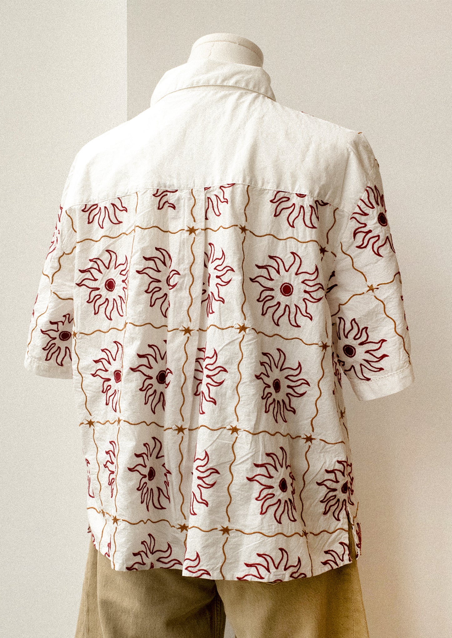 A short sleeve, collared shirt in white with red and brown sun embroidery.