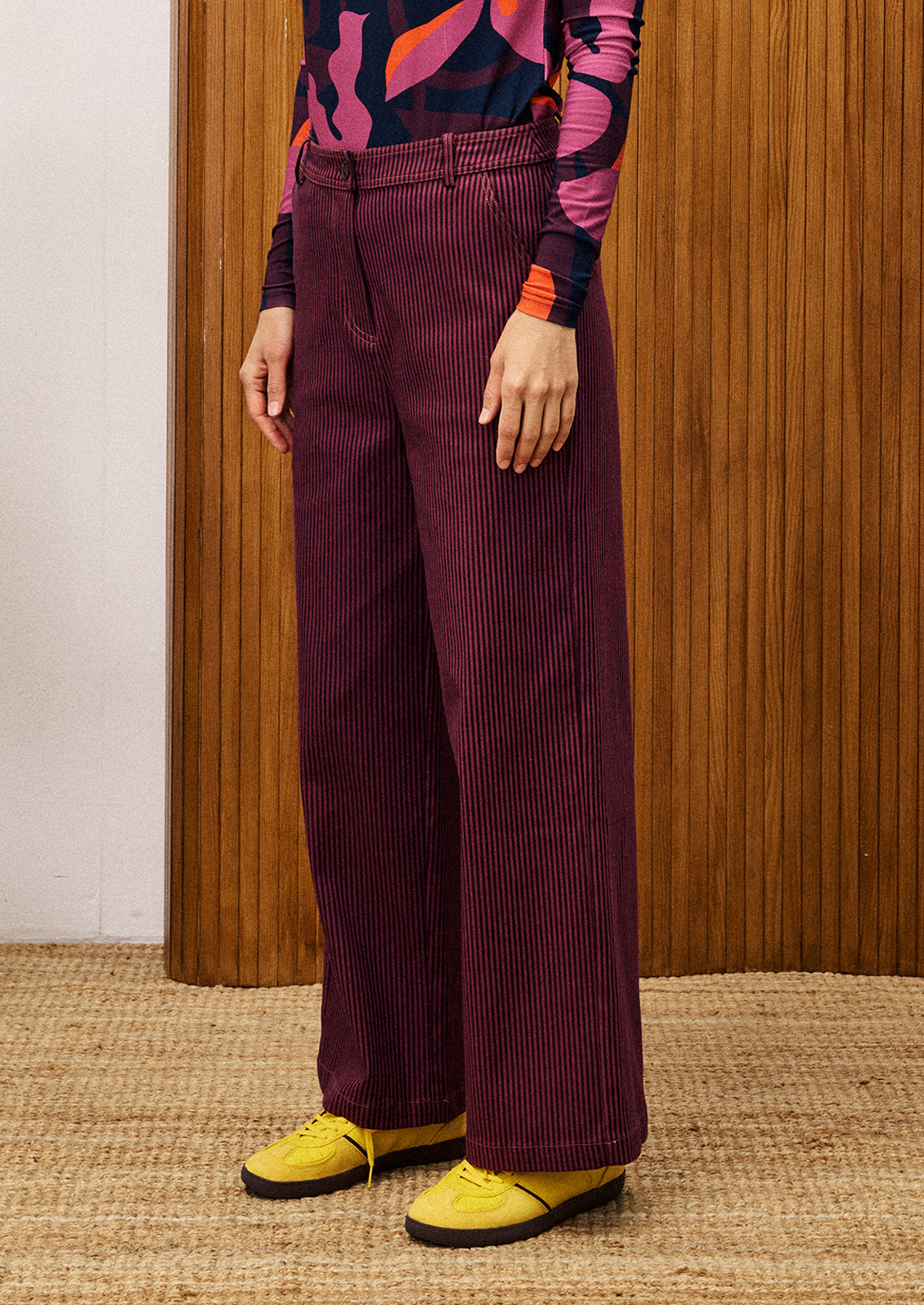Person wearing a colorful top and purple corduroy pants with yellow shoes, standing against a wooden panel background.