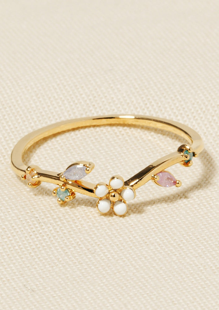 A gold ring with pastel colored crystals in mixed shapes, white enamel flower at middle.
