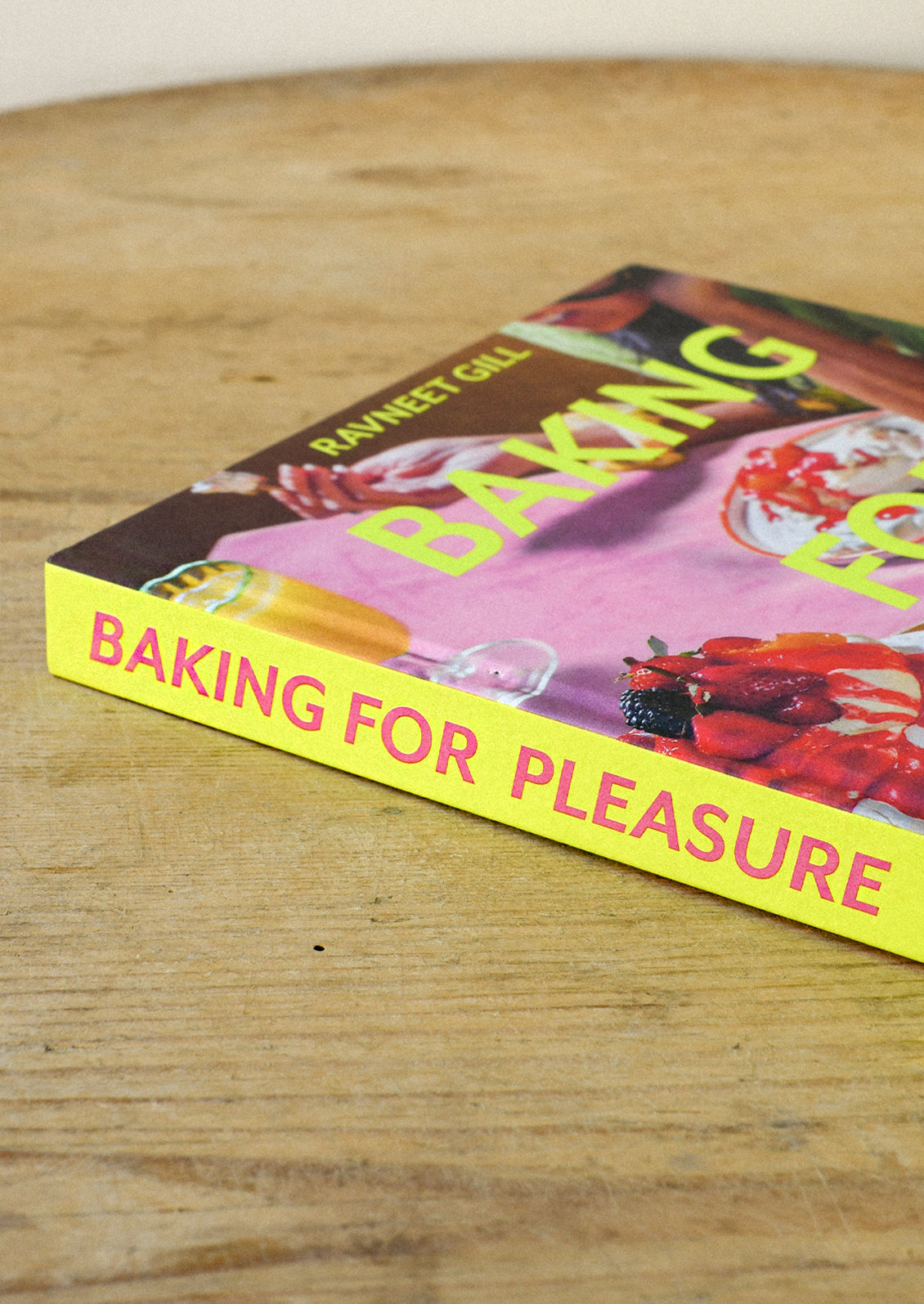 A cookbook titled "Baking for Pleasure" by Ravneet Gill on a wooden table.