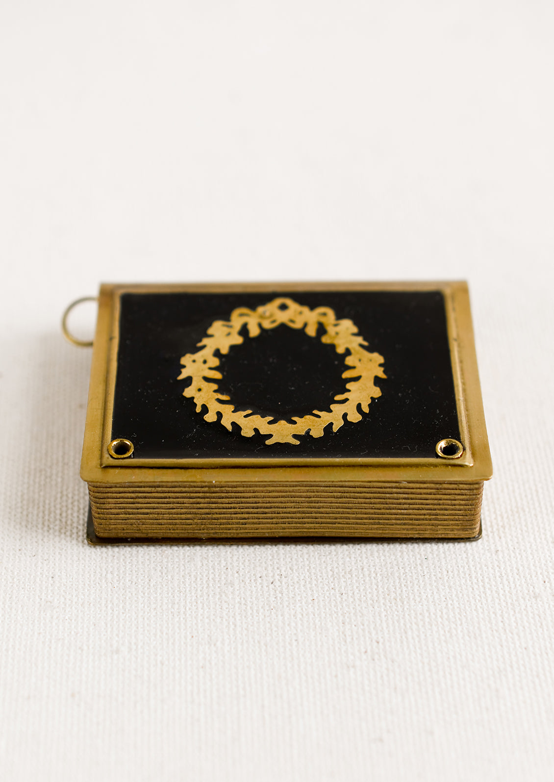 A book-shaped measuring tape with brass wreath design on black enamel.