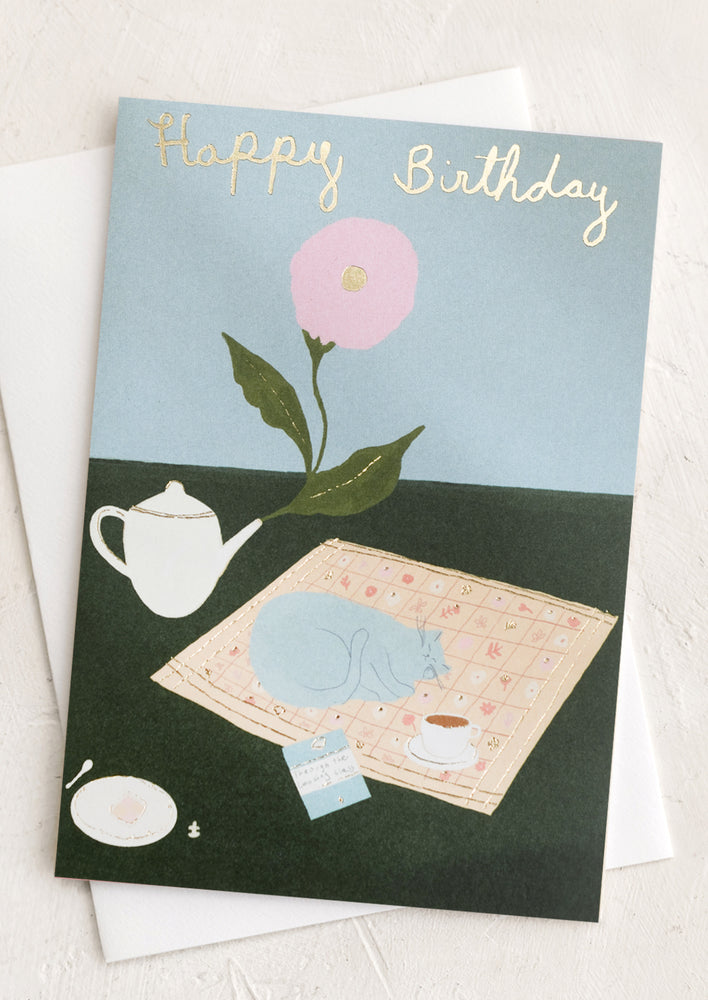 A card with illustration of cat napping on a picnic blanket, text reads "Happy Birthday".