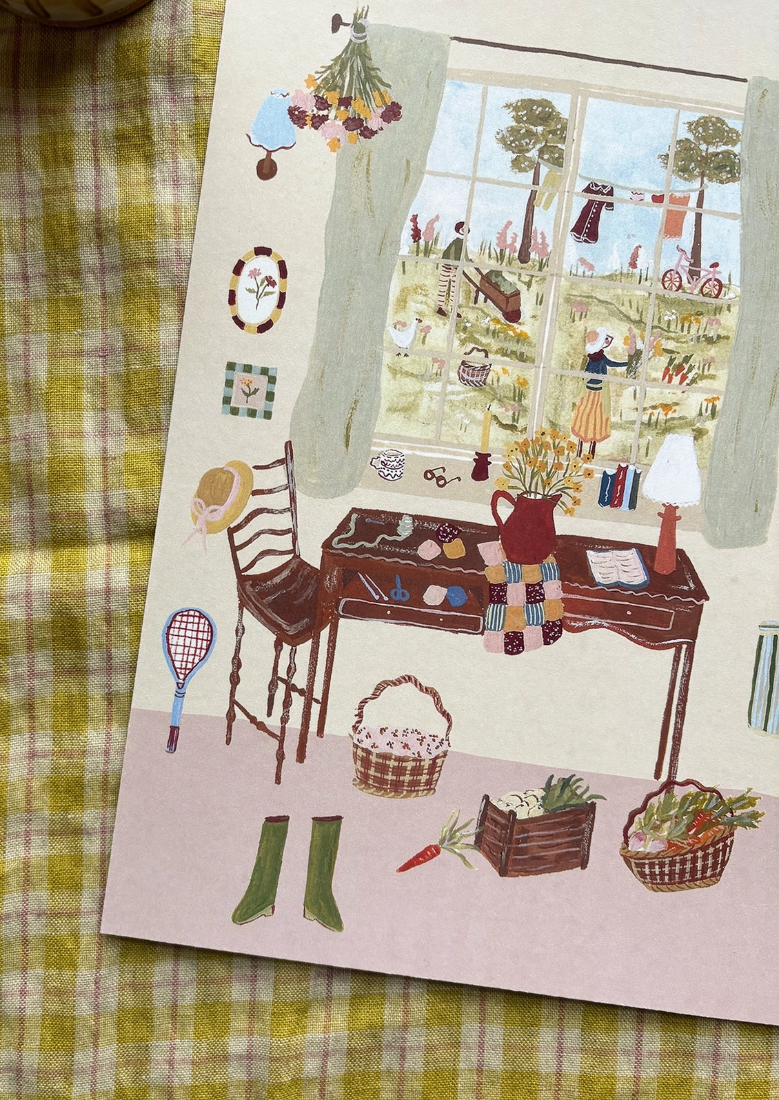 An art print of an illustration of a countryside setting, quilting supplies at a table next to a window.