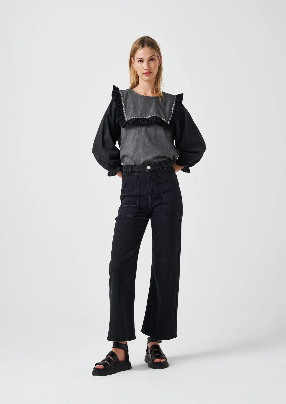 A woman standing wearing a black top with black jeans and black sandals.