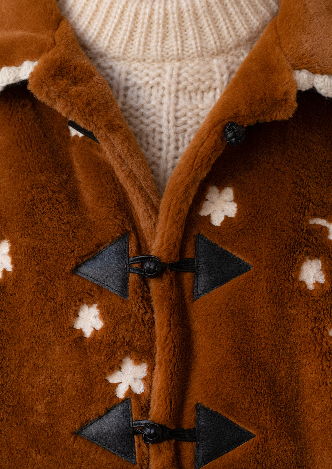A woman wearing a caramel colored faux fur jacket with white floral embroidery and black toggle button front.