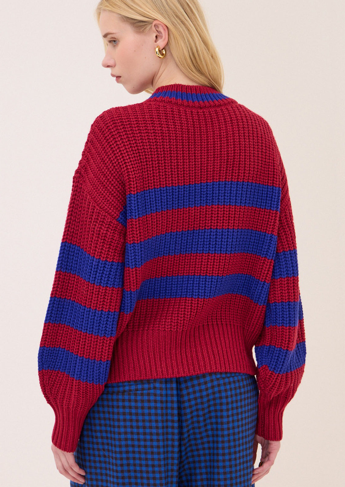 A woman shown from the back wearing a chunky knit sweater with bold horizontal red and blue stripes, paired with blue and black checkered pants, against a plain light beige background.