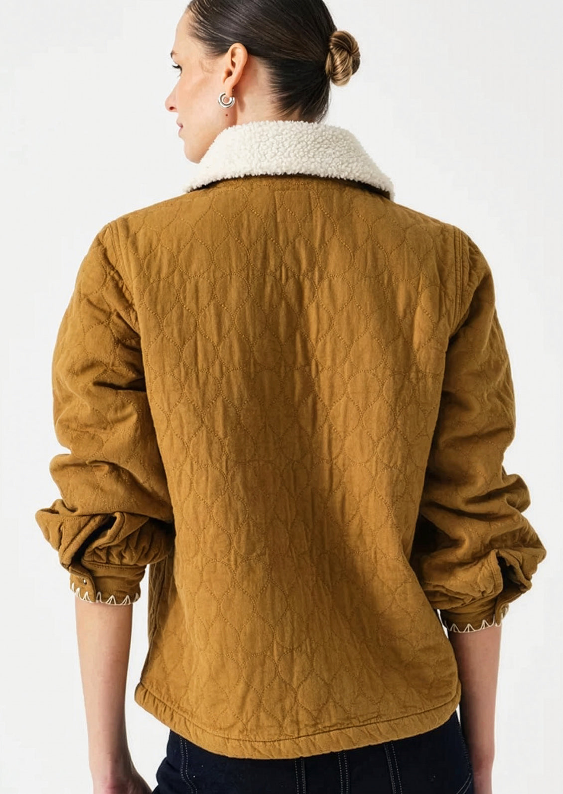 A woman shown from the back wearing a mustard yellow quilted jacket with a white sherpa collar, set against a plain white studio background.