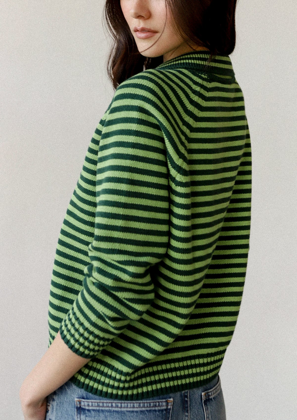 A woman wearing a green and dark green horizontally striped knit sweater, shown from a side-back angle against a plain light gray wall.