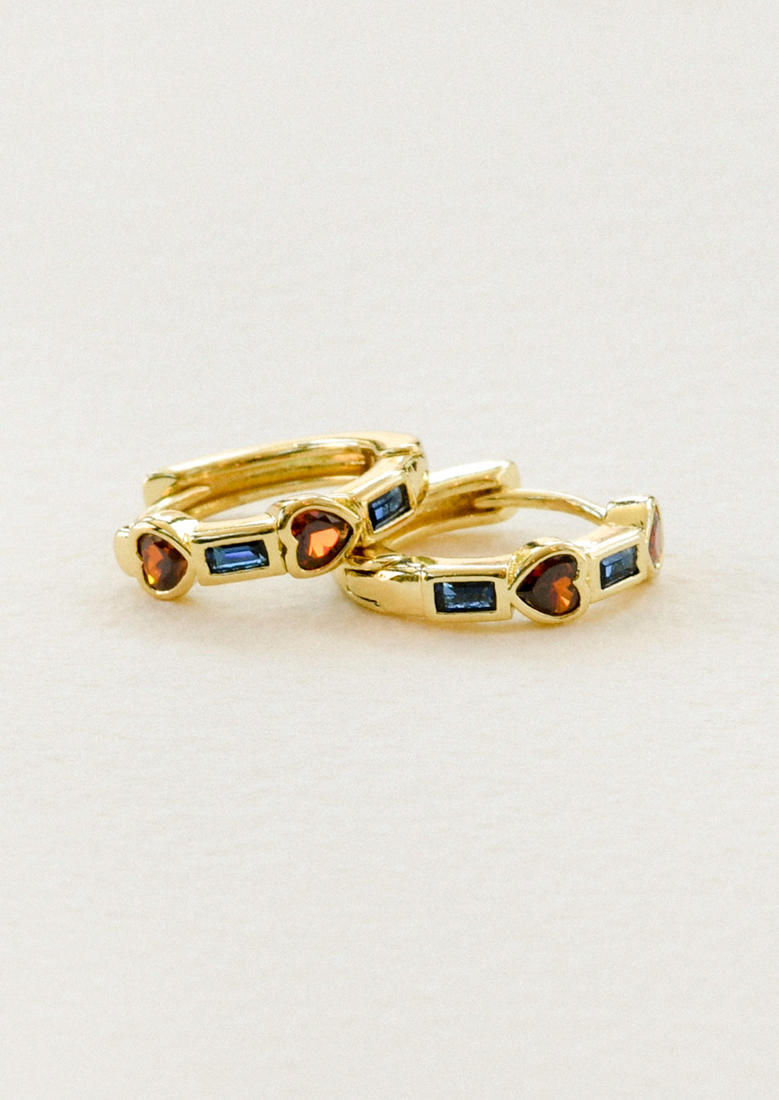 A pair of gold huggie hoop earrings with alternating red heart and blue rectangular inlaid gems.