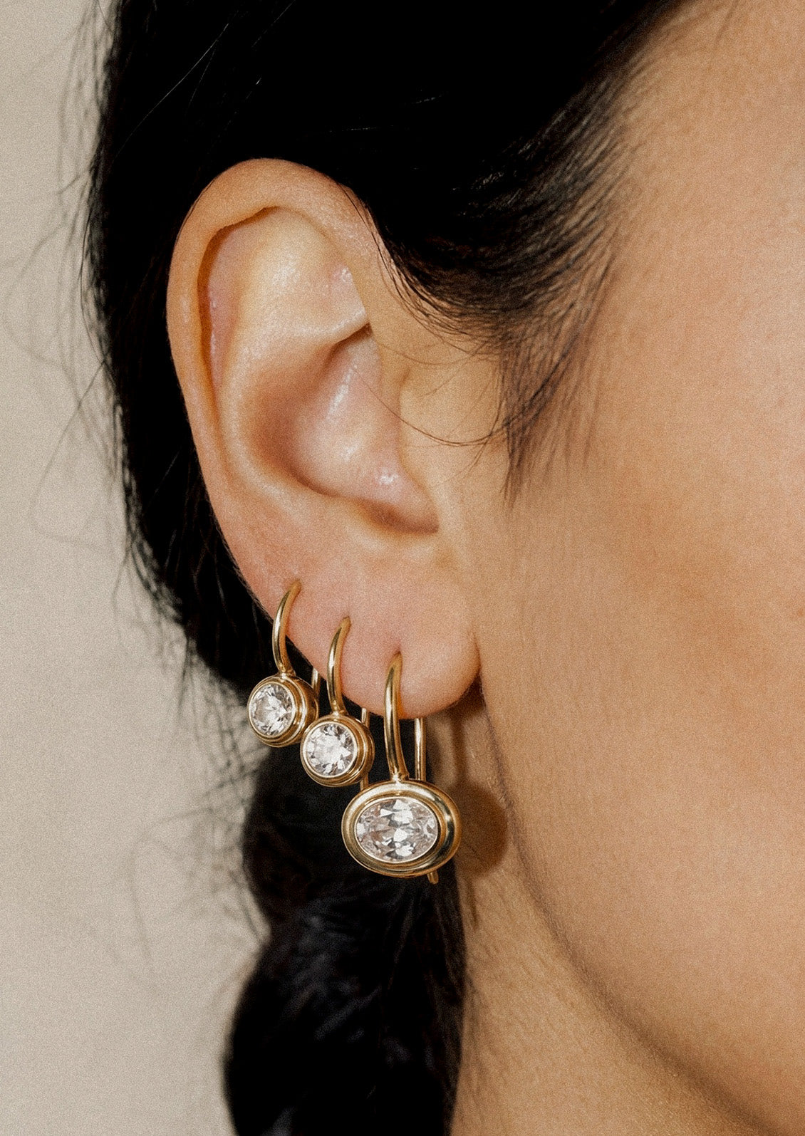 A woman wearing three gold dangling hook earrings with a clear lab grown sapphire in the center.