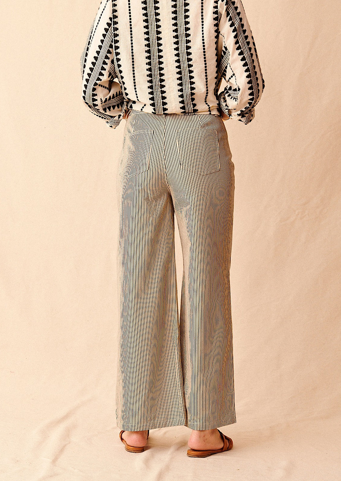 A woman standing wearing cream and black pinstriped wide leg jeans with brown sandals and cream and black embroidered top, seen from behind