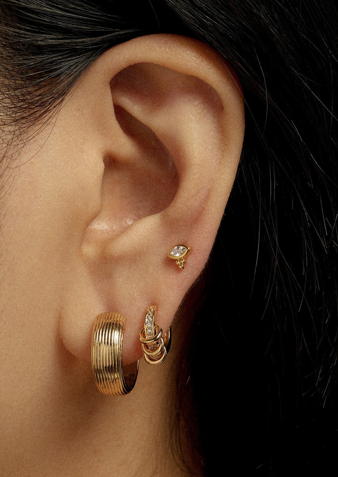 A woman wearing a variety of gold earrings on her ear including a stud earring and two hoop earrings.