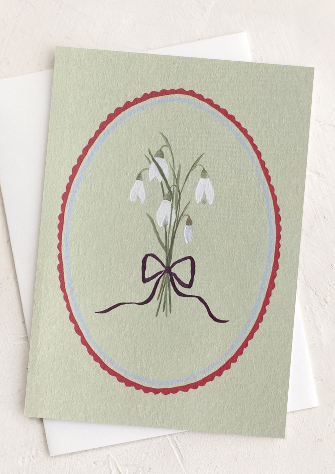 A sage green greeting card with oval frame around snowdrop flowers.