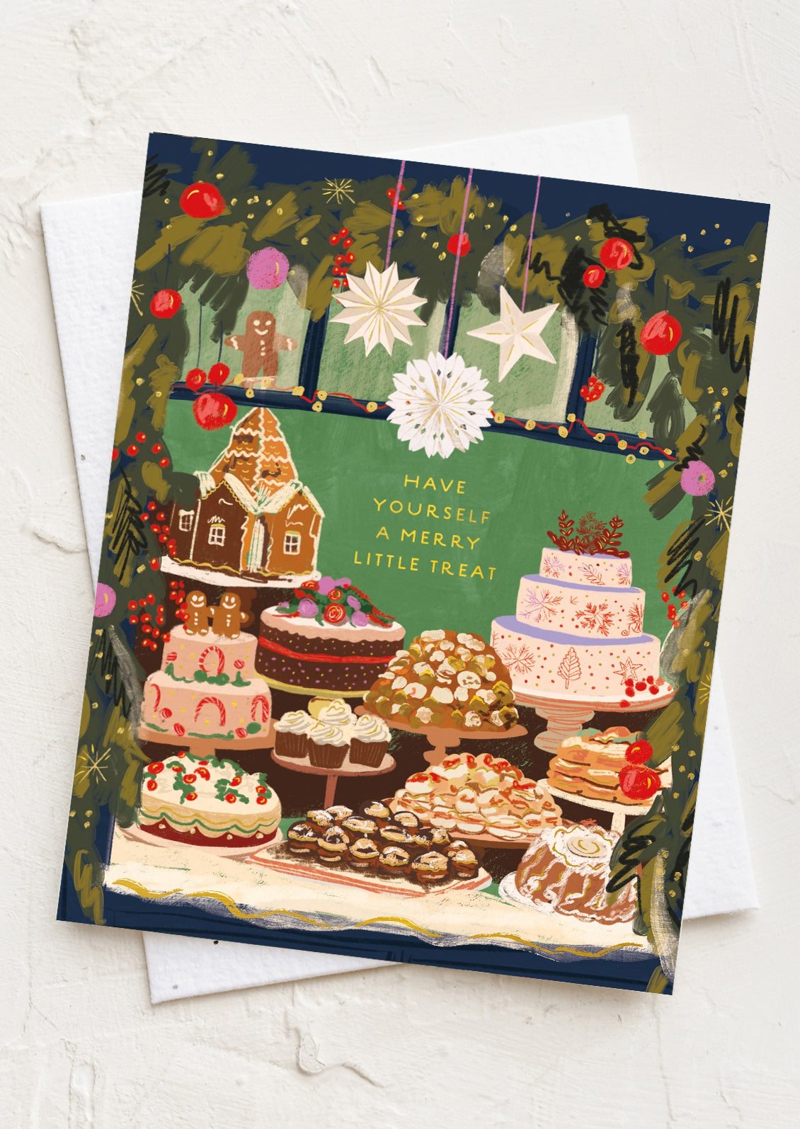 A Christmas-themed illustrated greeting card featuring an assortment of cakes, pastries, and a gingerbread house, with the text 'HAVE YOURSELF A MERRY LITTLE TREAT'. The card is displayed on top of a white envelope against a textured white background.