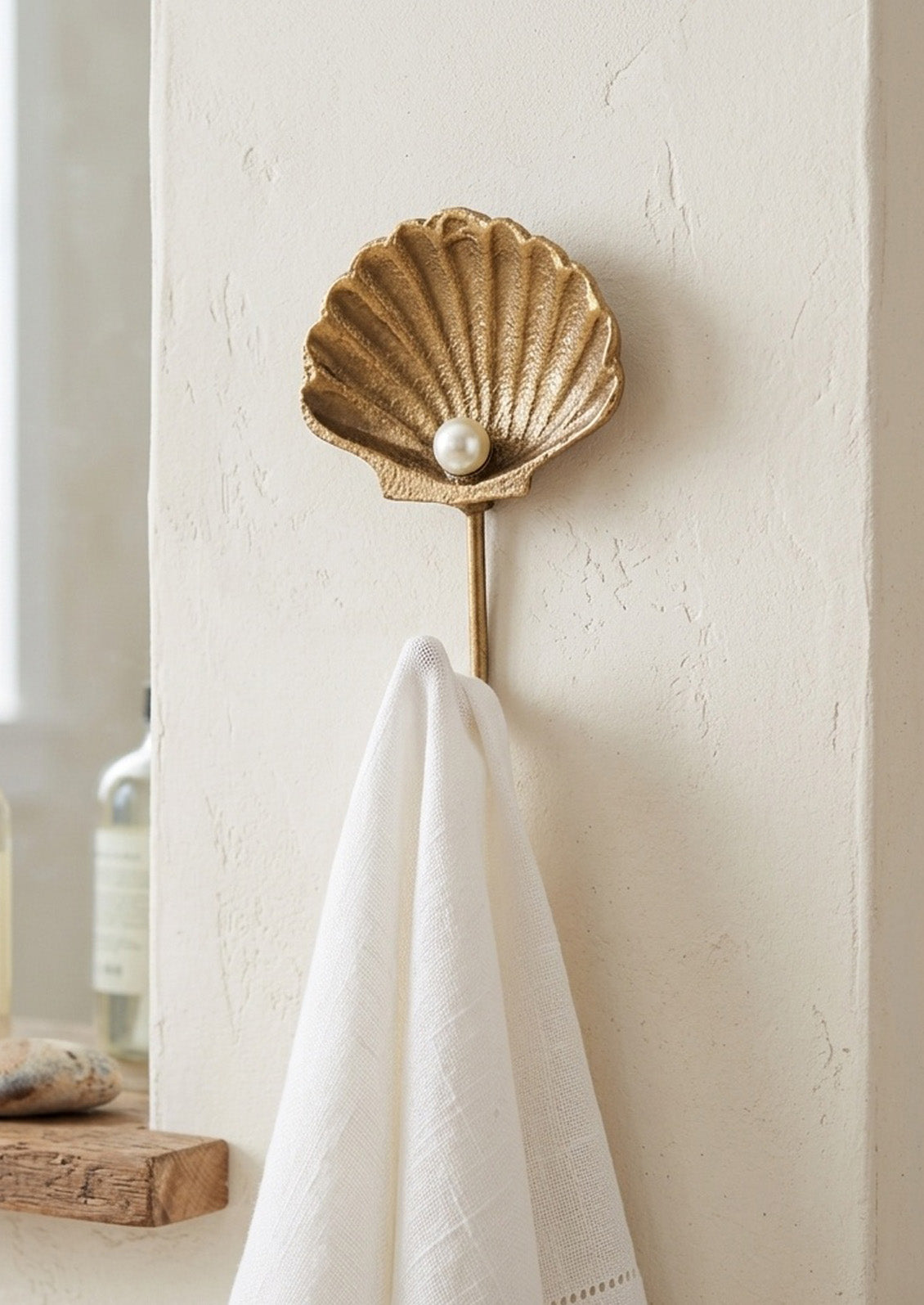 A handcrafted gold cast iron wall hook in the shape of a shell with a faux pearl in the center hanging on a white textured wall in a bathroom with a white linen hanging on the hook.