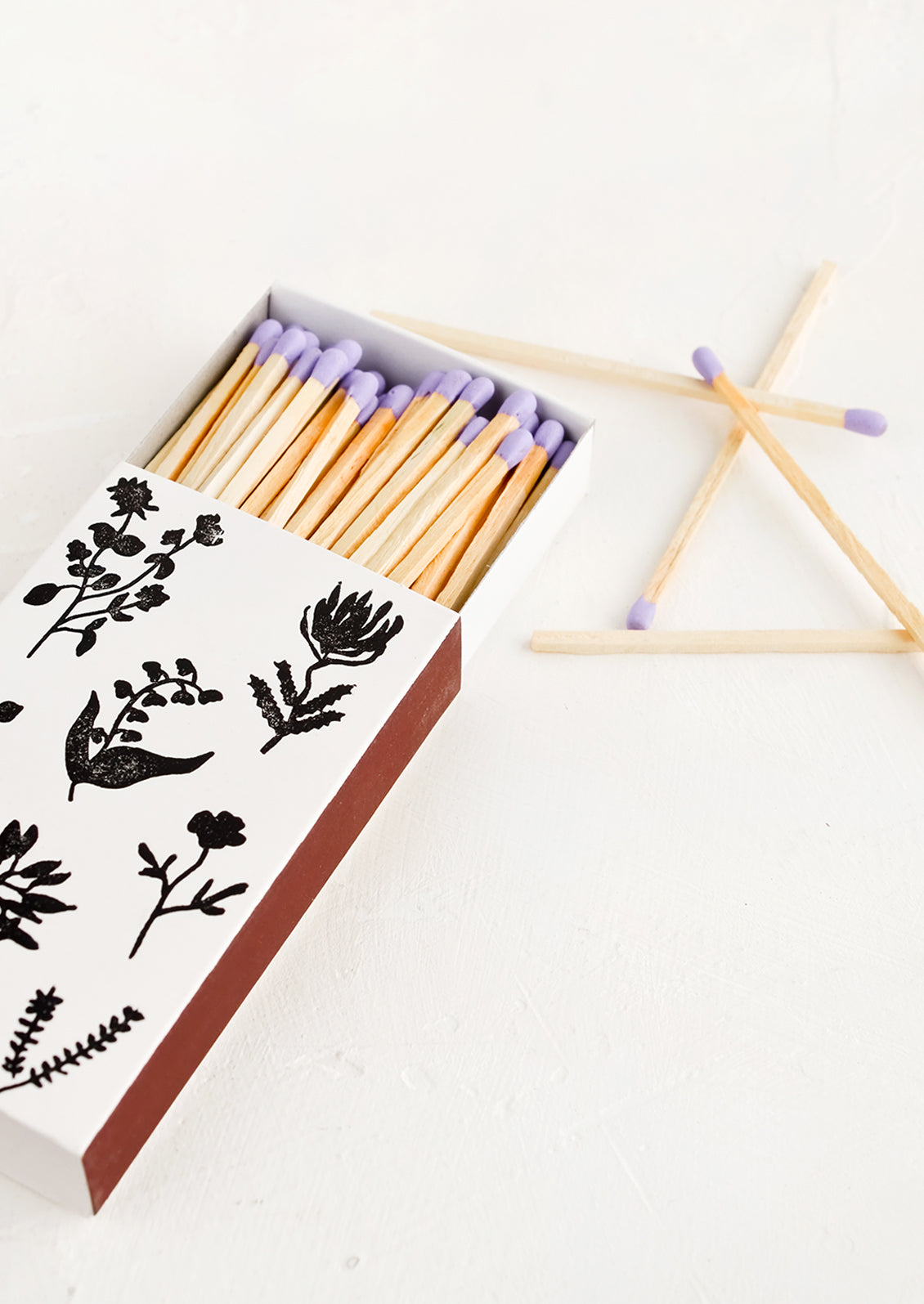 A matchbox in white with black botanical print, housing purple tipped matches.