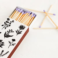 3: A matchbox in white with black botanical print, housing purple tipped matches.