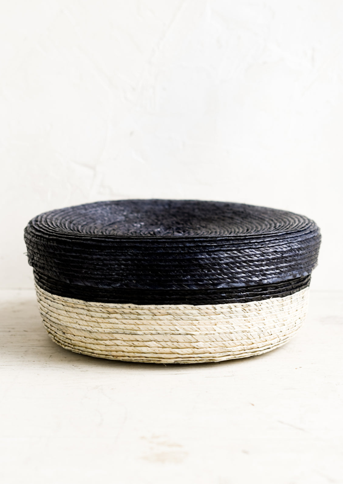A shallow, round storage basket with lid, woven from dried palm leaf in two-tone design.