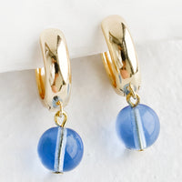Sky Blue: A pair of gold huggie hoop earrings with single spherical blue glass bead detail.