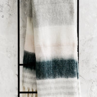 Pink Multi: A striped throw blanket in pink and grey with fringe trim on a black ladder.