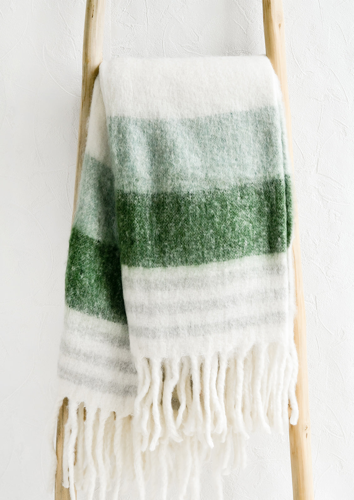 A striped throw blanket in green and grey with fringe trim on a wooden ladder.