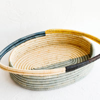 1: Oval bread basket in woven raffia with protruding handles at either side. Blue and yellow colorblock pattern.