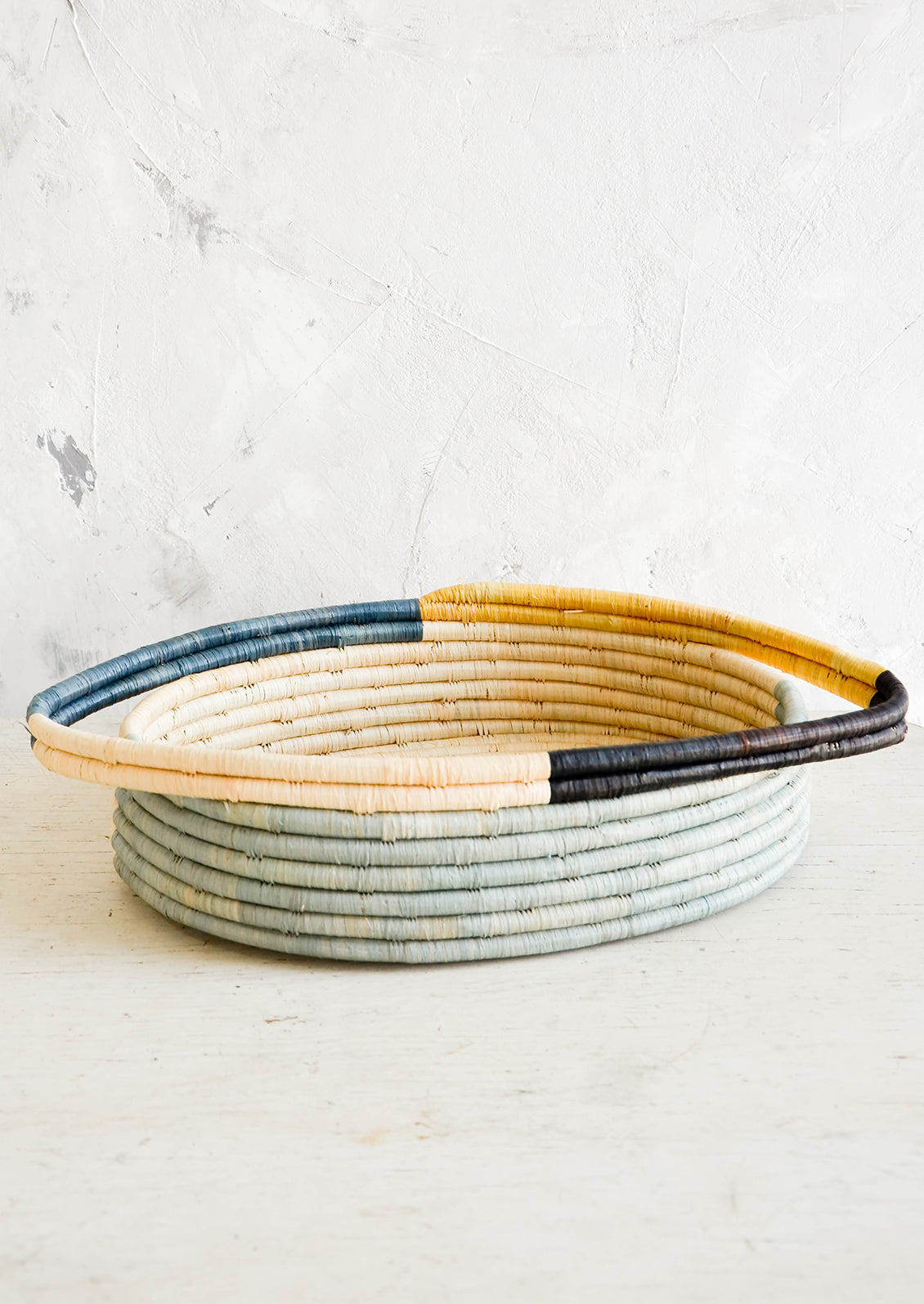 Oval bread basket in woven raffia with protruding handles at either side, sitting on a table.