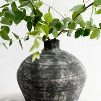 1: A distressed black clay vase with narrow opening, with eucalyptus branches.