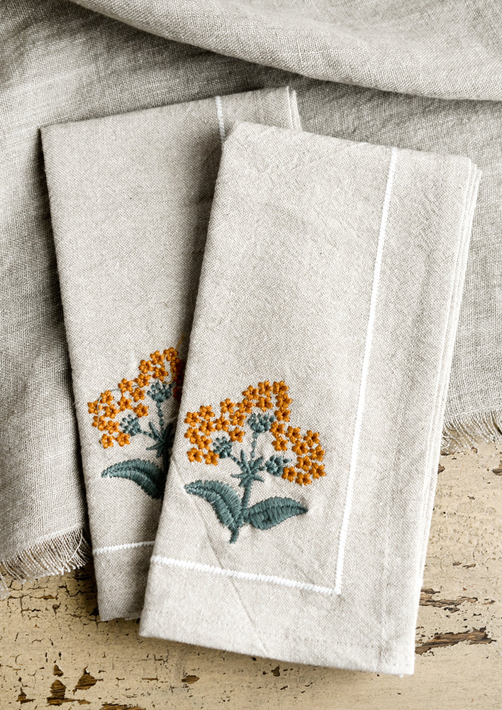 A pair of natural cloth napkins with amber floral embroidery.