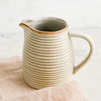 3: A tan ceramic pitcher with striped texture on top of pink linen.