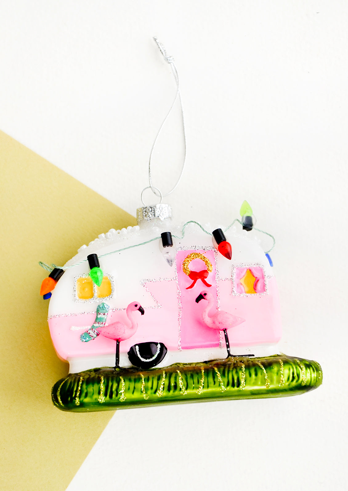 A glass ornament in the shape of an RV decorated for Christmas with two flamingos out front.