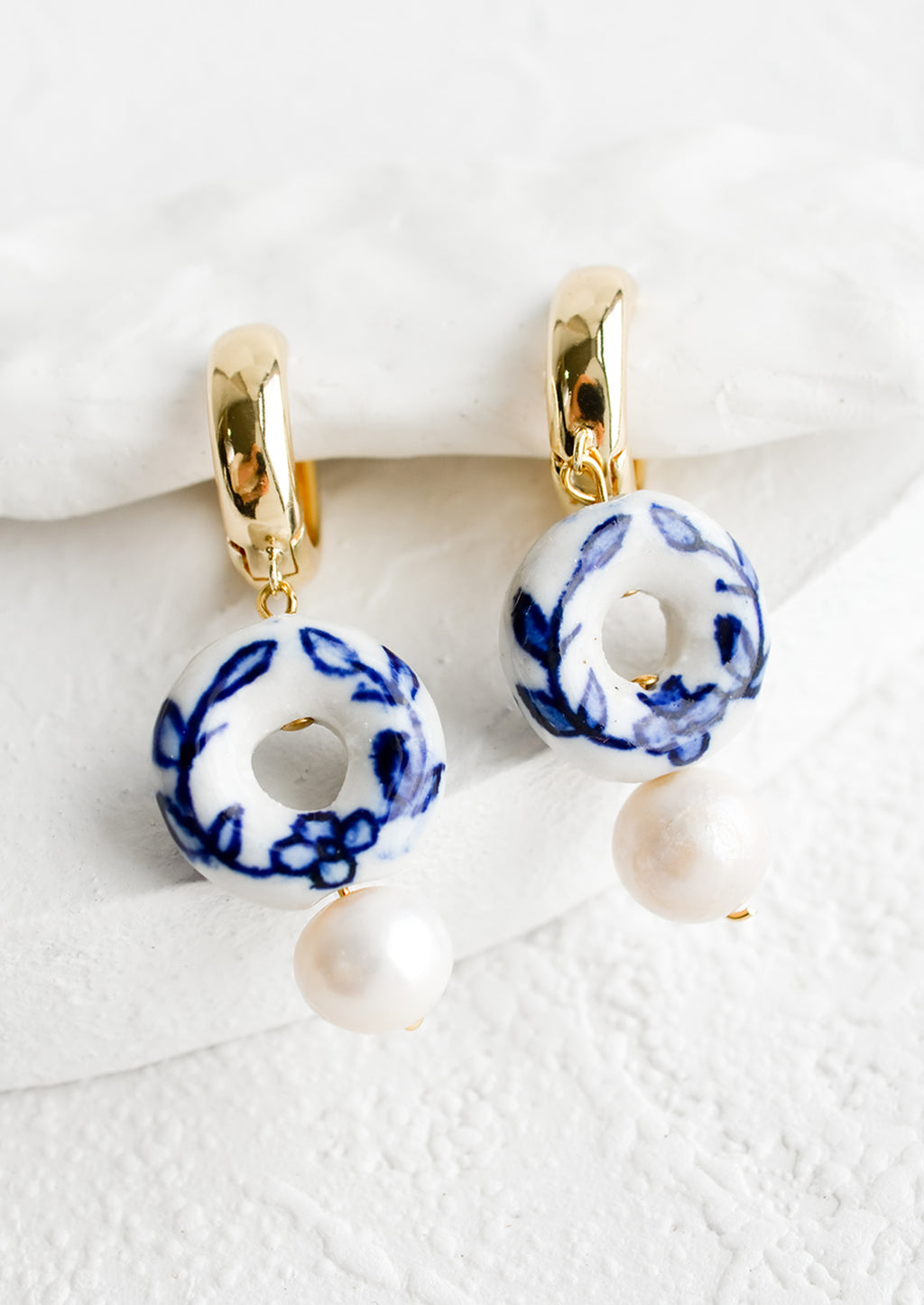 2: A pair of gold huggie hoops with donut shaped blue and white ceramic bead and pearl below.
