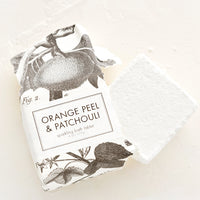 Orange Peel & Patchouli: White bath fizz cube emerging from black and white botanical packaging.