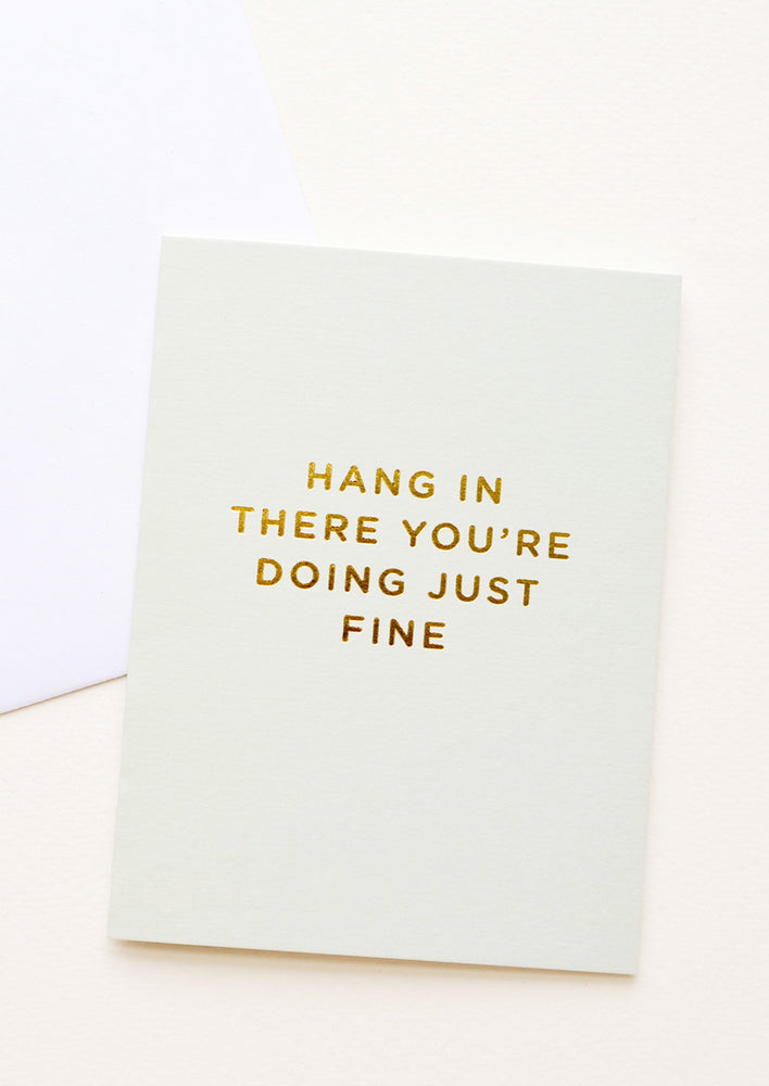 A pale green greeting card with the phrase "hang in there you're doing just fine" in gold foil.