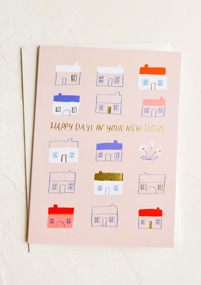 A pink greeting card with illustrated red and blue houses, text reads "Happy days in your new home".