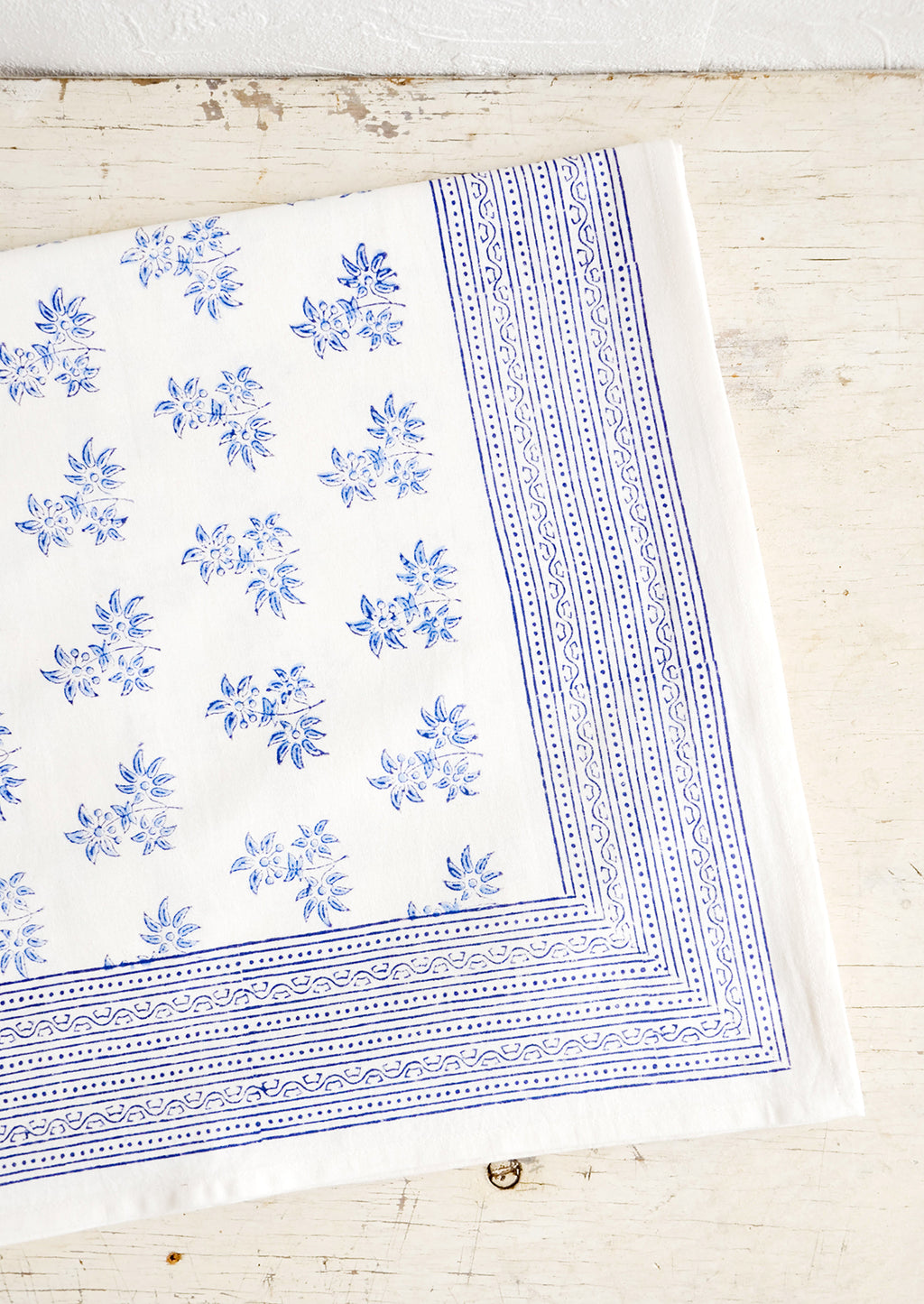 1: White cotton tablecloth with blue floral print, folded on an antique table.