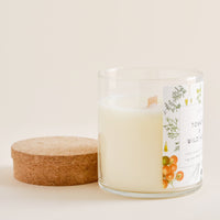 3: A glass candle with botanical label and wooden wick sits next to its cork lid.