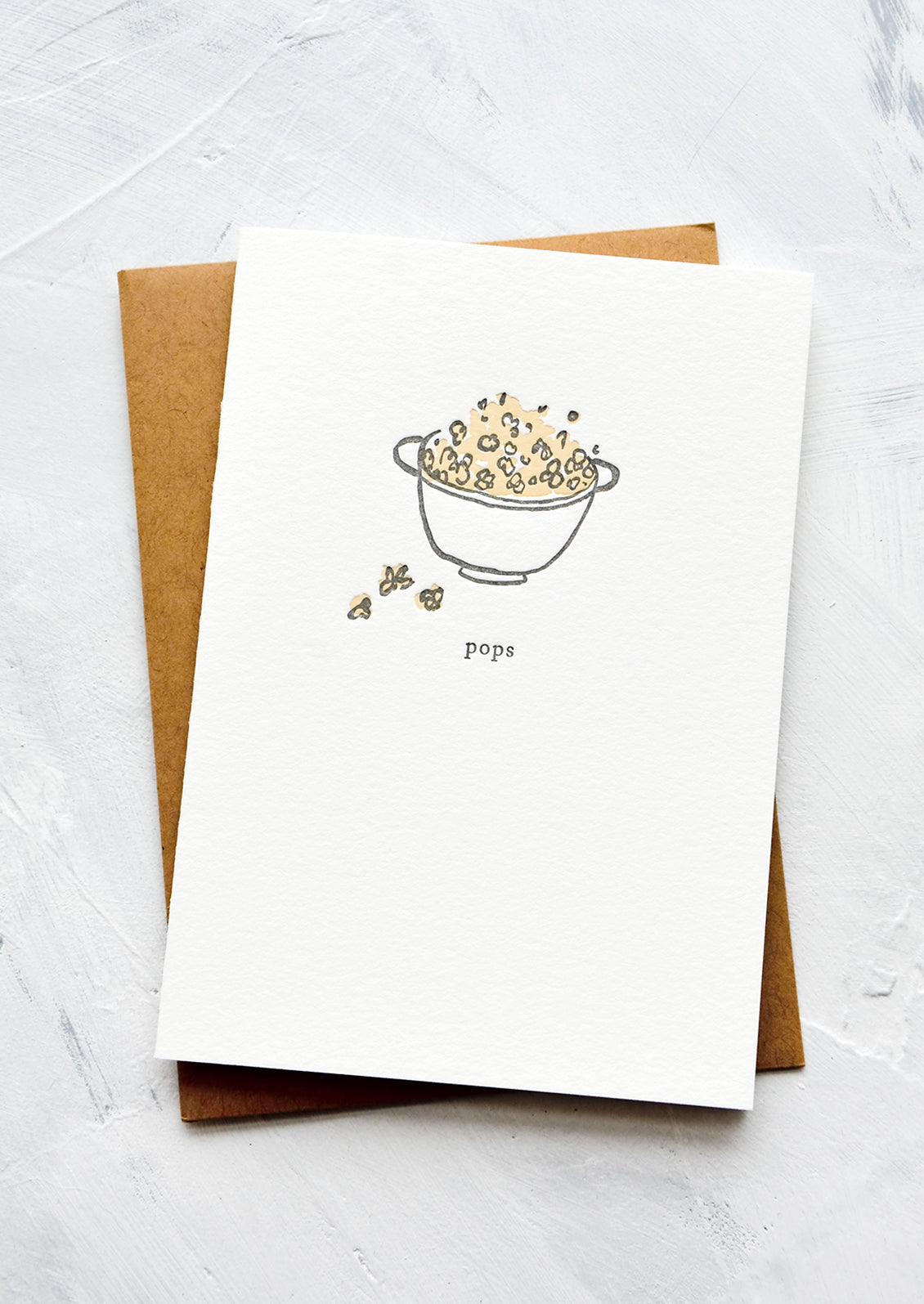 A letterpress printed greeting card with an image of a bowl of popcorn with text below reading "pops".