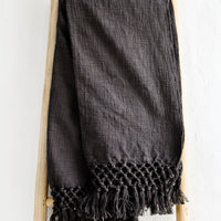 Carbon: A dark grey cotton throw blanket with knotted open weave trim, displayed on a ladder.