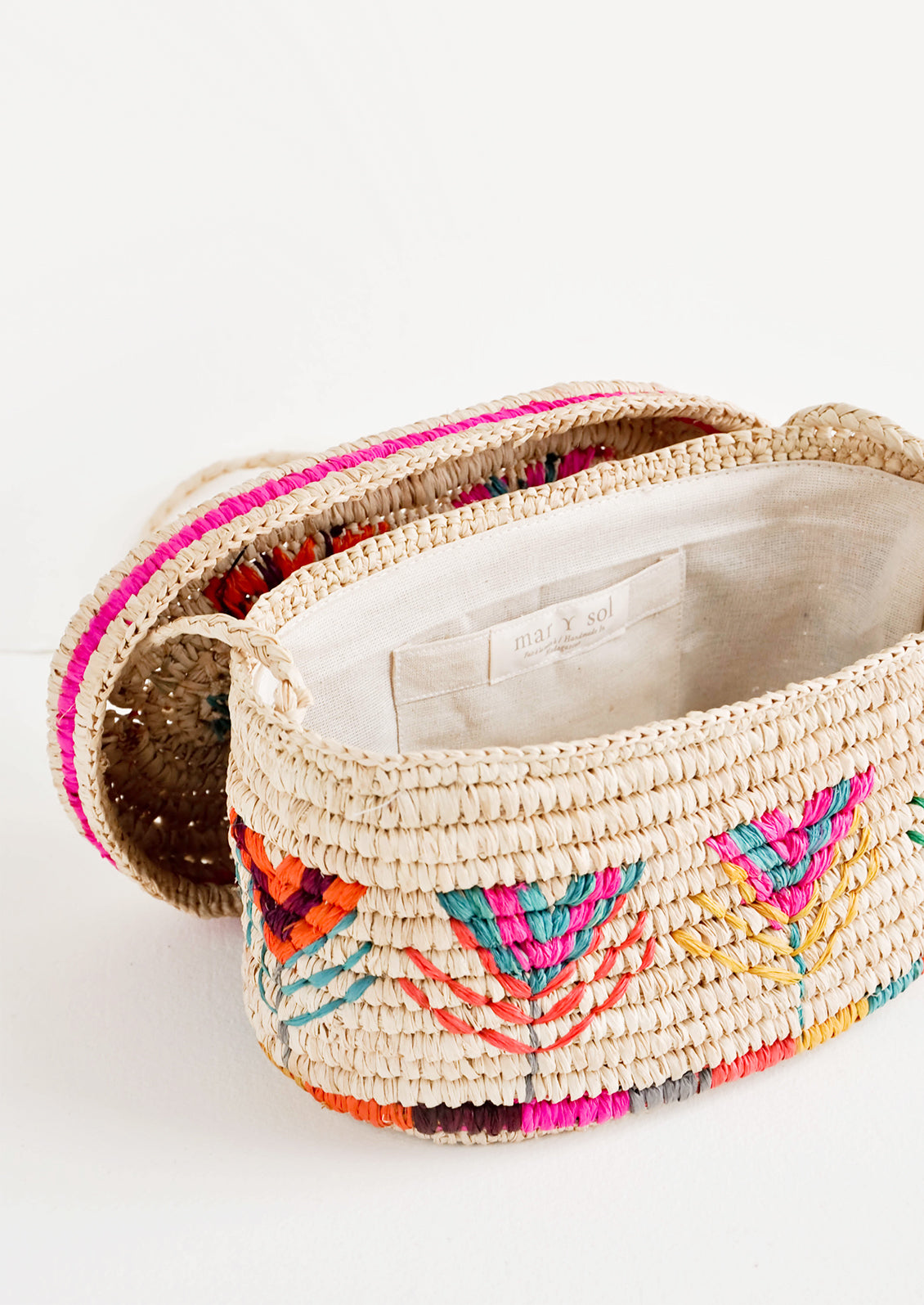 Rounded box style straw bag with neon multicolored flower pattern shown with lid off exposing cotton interior lining.