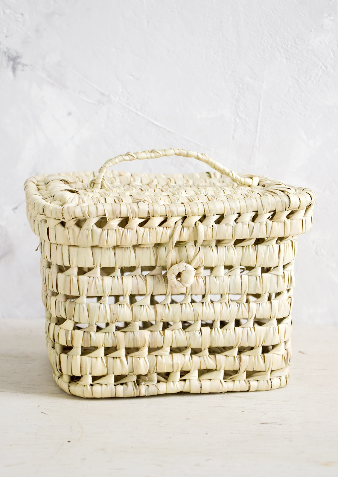 A square-shaped, lidded basket made from woven natural palm leaf.