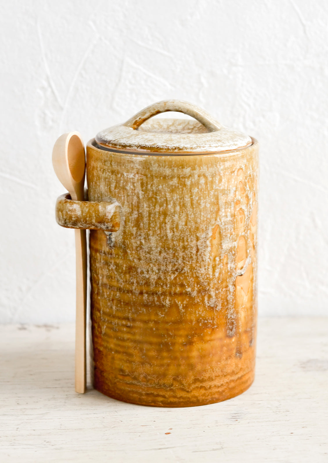 A ceramic storage jar in rustic light brown glaze with a wooden spoon on the side.