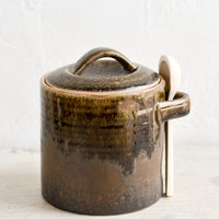 Short / Dark Brown: A ceramic storage jar in a rustic dark brown glaze with a wooden spoon on the side.